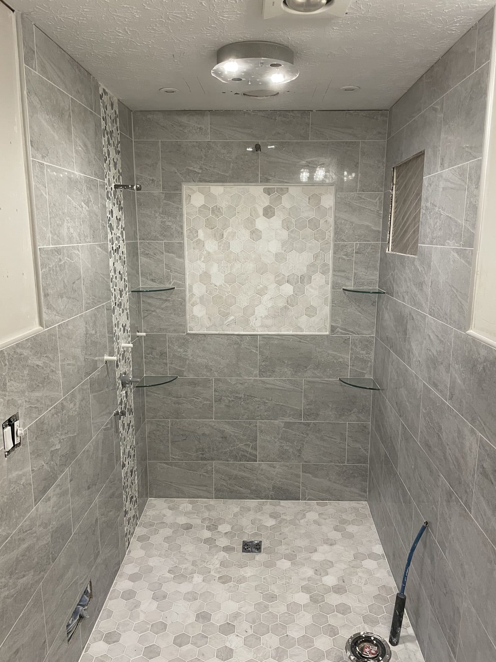 Large walk-in shower with gray stone tile, hexagon mosaic feature wall, pebble strip accent, and multiple glass shelves in Northeast Ohio